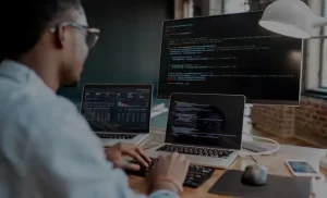 Person working at a desk with three screens displaying code and data analytics dashboards in a modern office setting