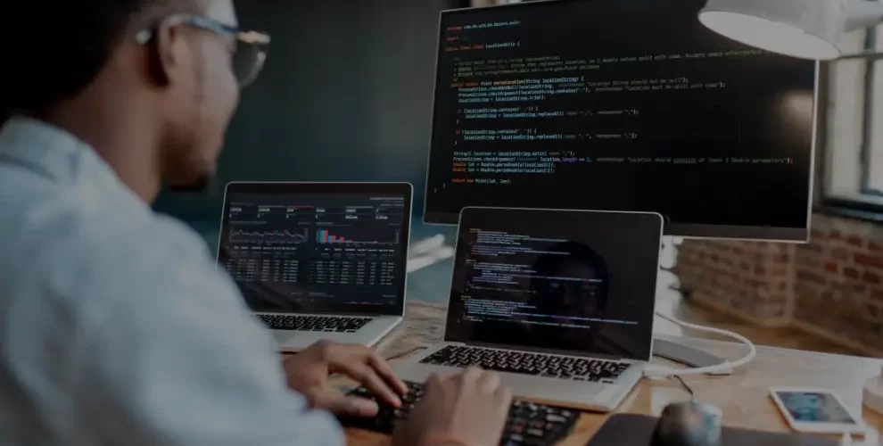 The Future Is Now: Exploring Low-Code No-Code App Development 1 Person working at a desk with three screens displaying code and data analytics dashboards in a modern office setting